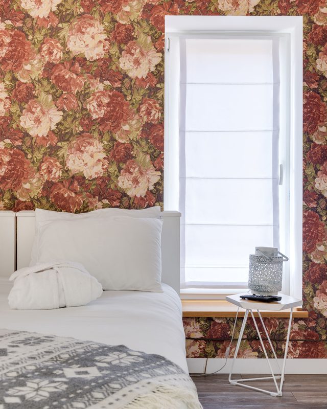 Bedroom with warm rose floral wallpaper beside a bright window with light bedding.