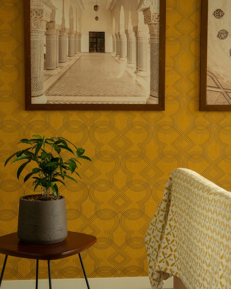 Warm mustard-gold geometric wallpaper on a living-room wall beside a potted plant and an upholstered chair.