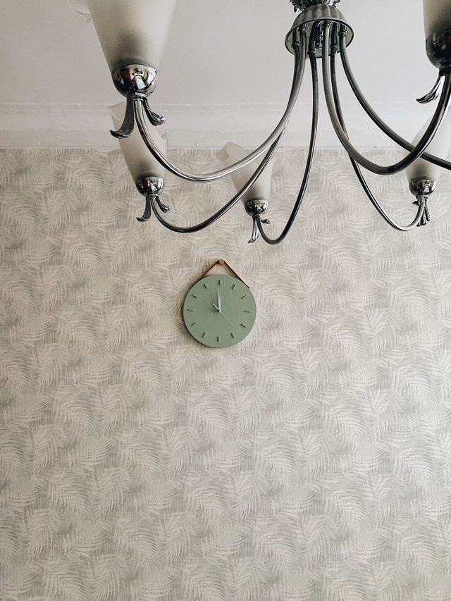 Softly patterned sage-green wallpaper in a light room with a wrought-iron chandelier and a round wall clock.