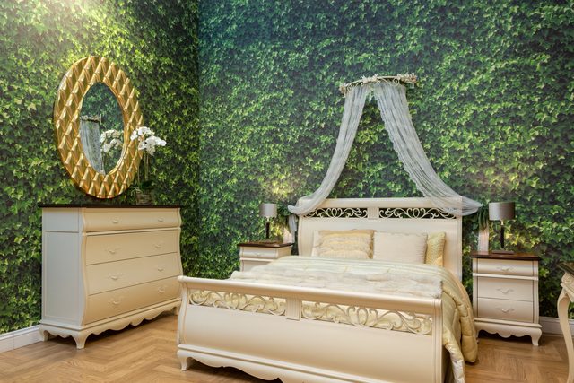 Realistic green ivy and leaf mural wallpaper wrapping two bedroom walls behind a carved cream bed and gold oval mirror.