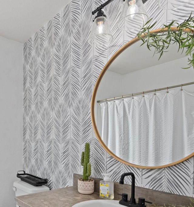 Powder room feature wall in a monochrome black-and-white palm-frond wallpaper with a round wood-framed mirror.