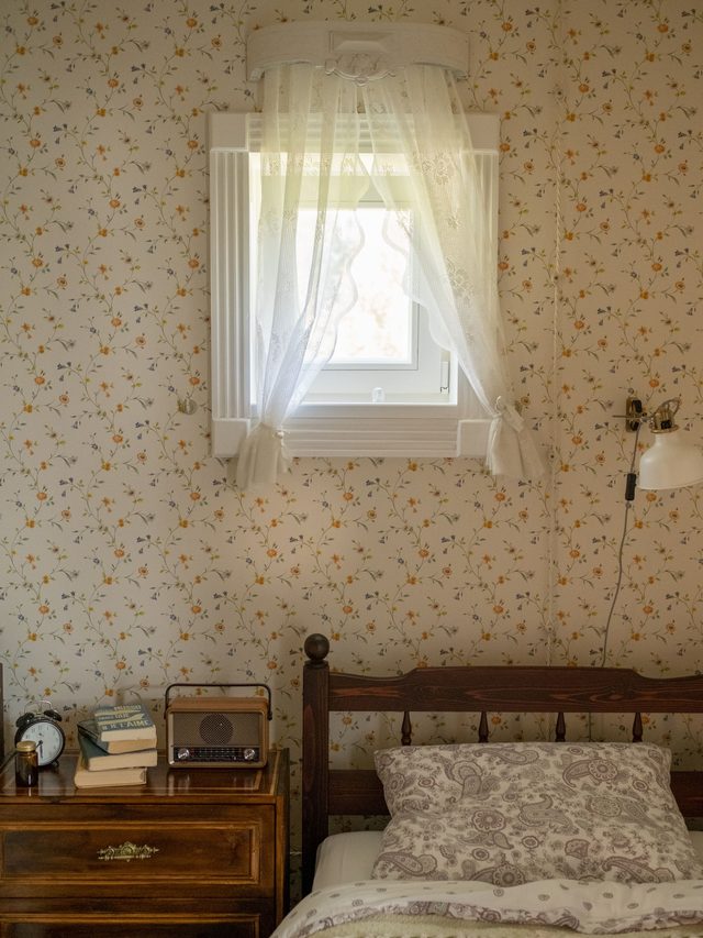 Cozy bedroom corner papered in a fine vintage floral pattern beside a curtained window and wooden bed.