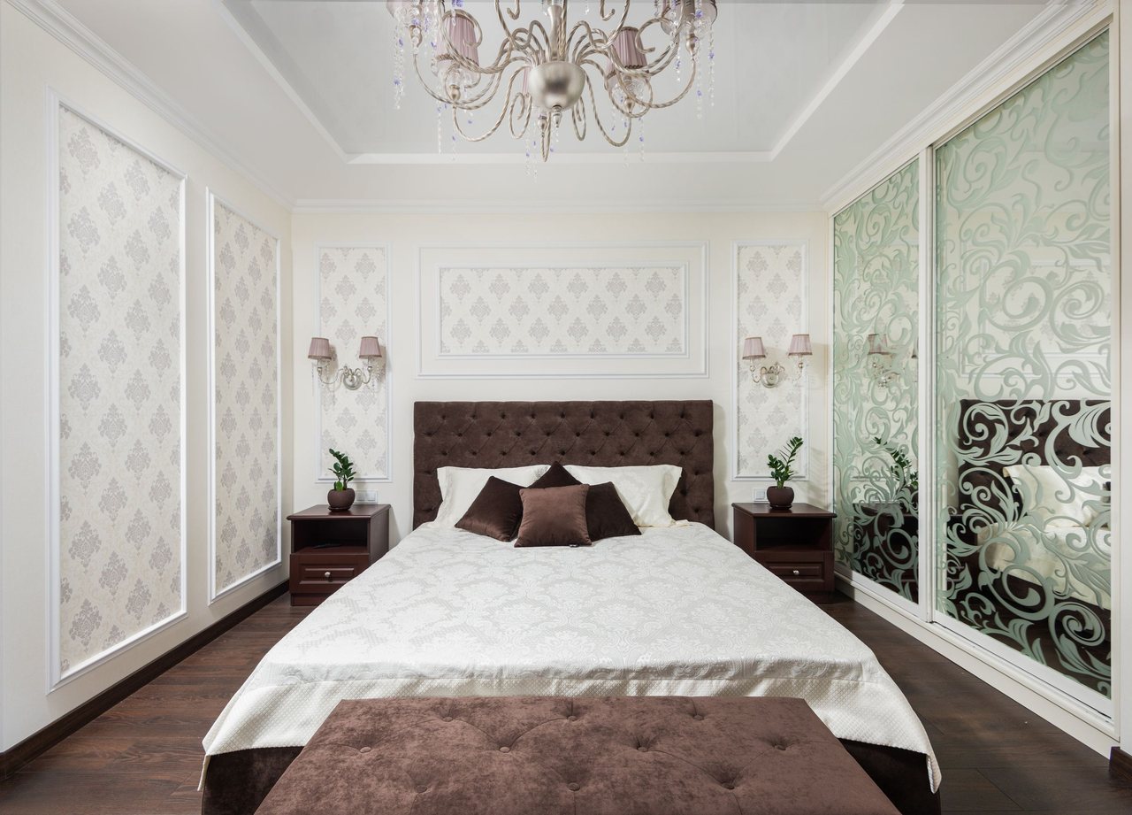 Elegant bedroom with cream and grey damask wallpaper set into moulded wainscot panels, a tufted headboard and a chandelier.