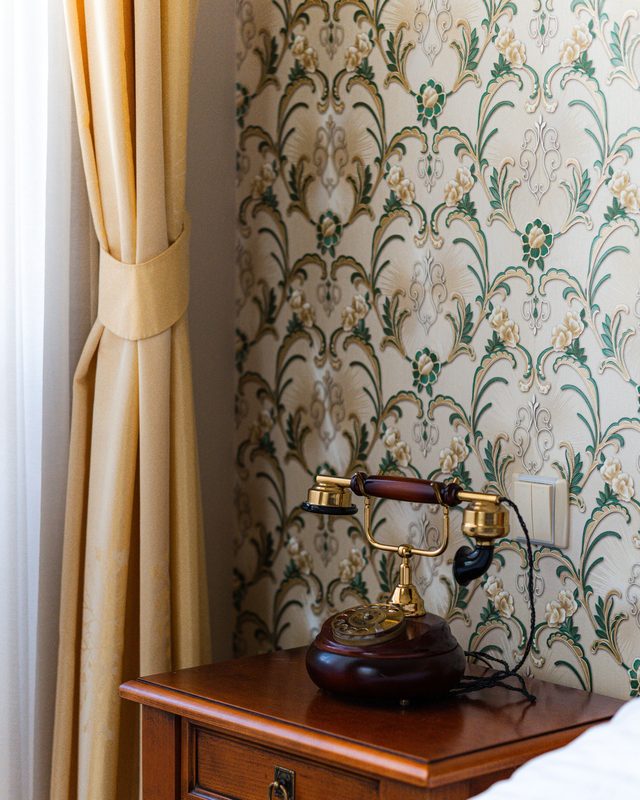 Green art-nouveau floral wallpaper on a wall beside a curtain and a vintage telephone on a wooden desk.